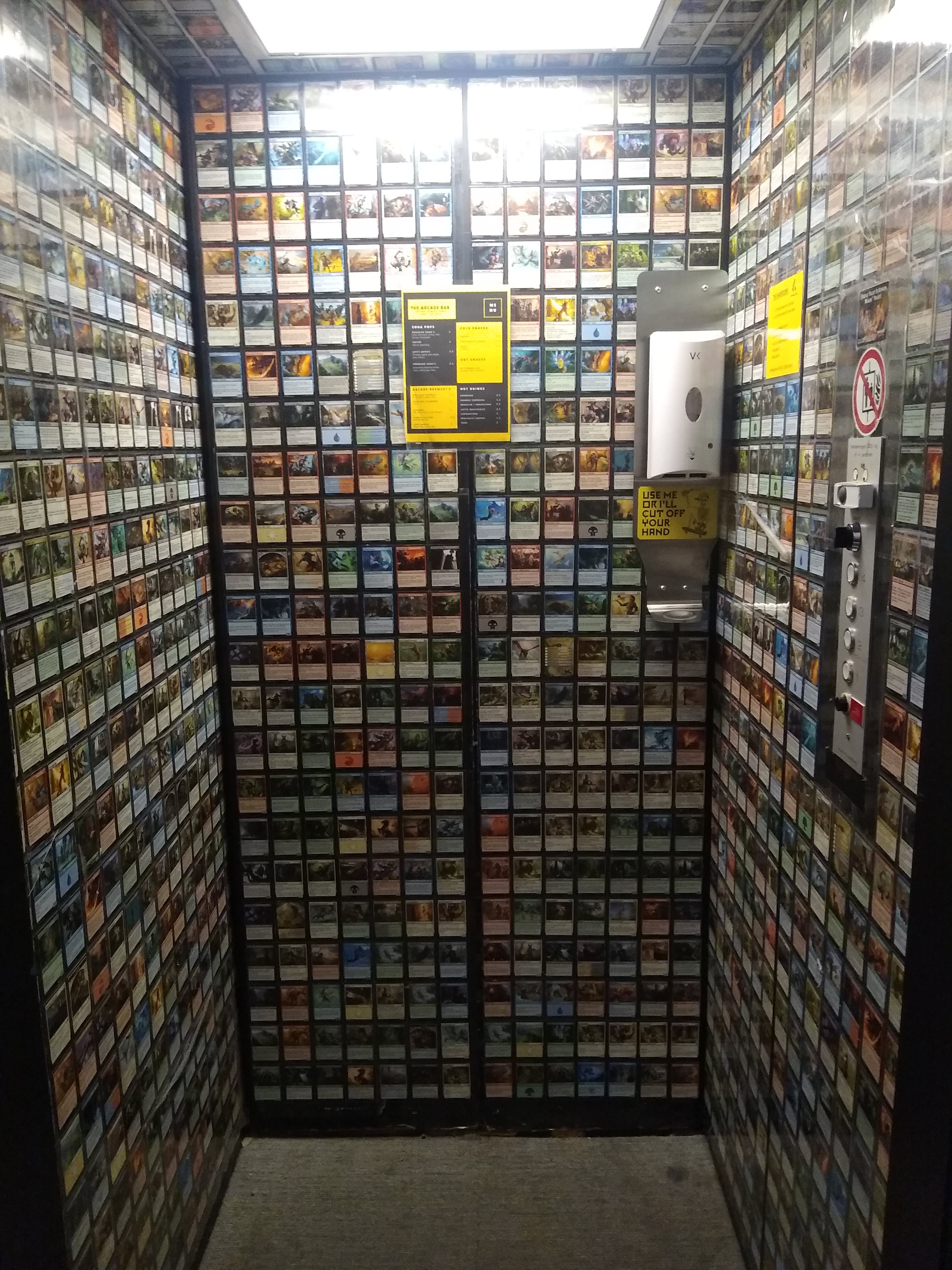 A photograph of the inside of a lift that is covered in Magic: The Gathering playing cards. The lift buttons are on the right. A white hand sanitiser dispenser is on the front wall. Some yellow signs featuring instructions for hand cleaning and a menu are placed on the walls.