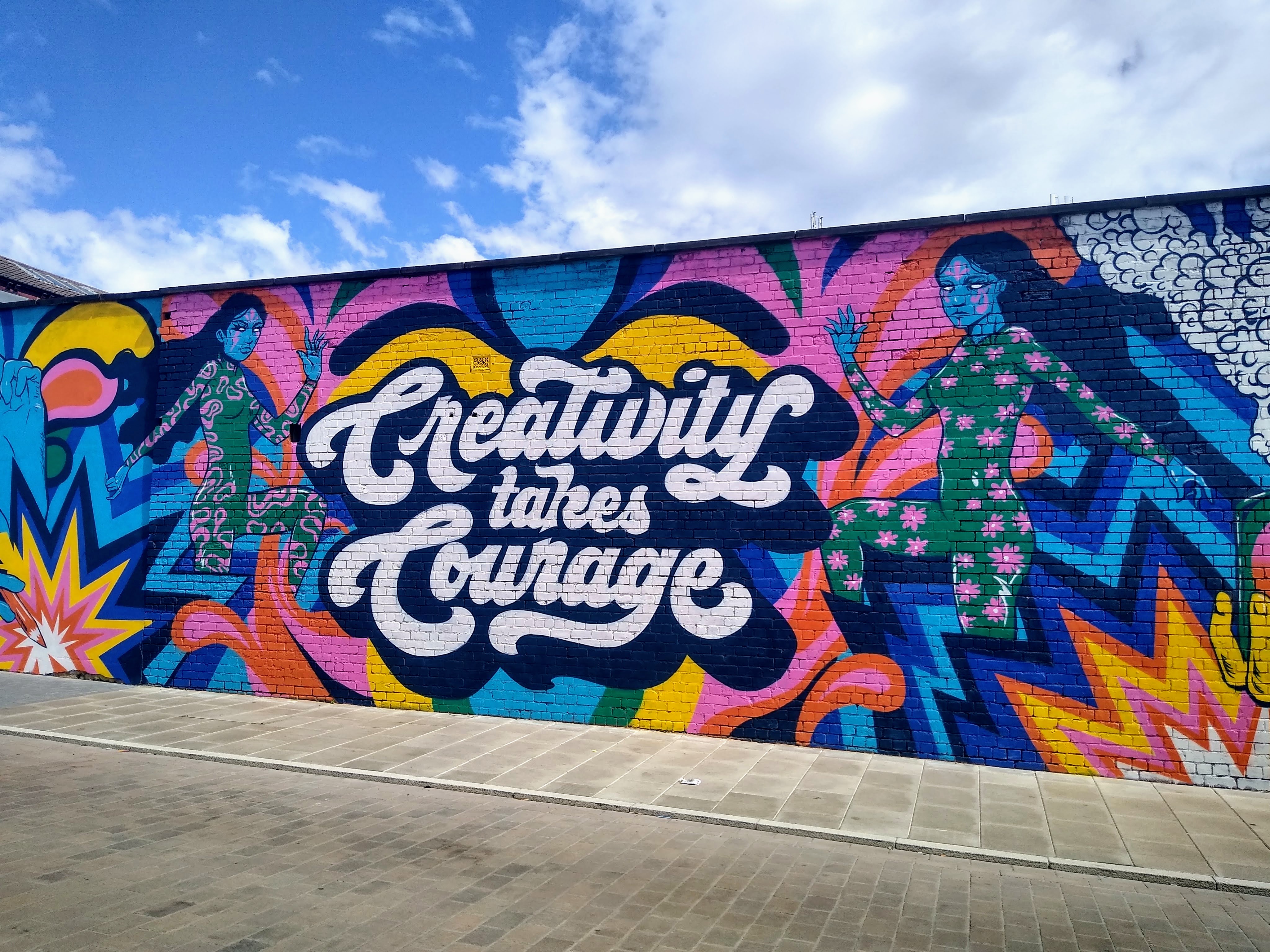 A photograph of a colourful mural on an urban wall. The mural depicts two female figures atop a background of waves and sharp-edged explosion-like imagery. Text in the middle, painted in a 60s style, reads 'Creativity Takes Courage'.